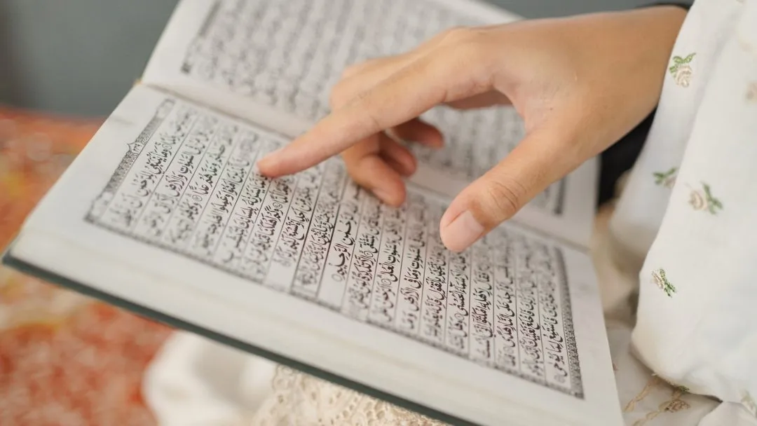 Close-up of a student's hand pointing to Arabic verses of the Holy Quran while studying through an interactive Islamic education program online.